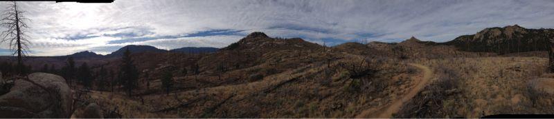 Panoramic view of a rugged landscape with rolling hills, sparse vegetation, and a winding dirt trail. The sky is filled with clouds, creating a dramatic backdrop over the mountainous terrain. Buffalo Creek mountain bike trail.