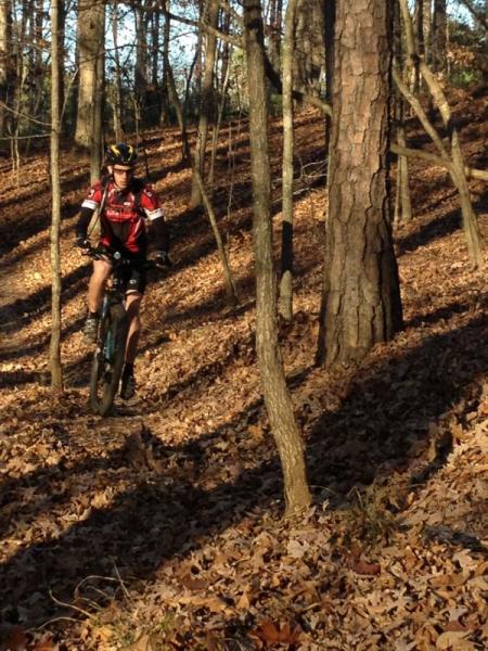 A mountain biker navigating a wooded trail surrounded by tall trees and fallen leaves in a sunlit forest. The cyclist is wearing a red and black jersey and a helmet, showcasing an active outdoor lifestyle. Blankets Creek mountain bike trail.