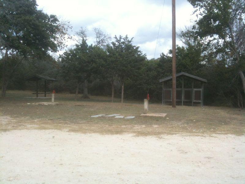 A grassy area with two wooden shelters and a picnic table, surrounded by trees. There is a utility pole in the background and a few concrete pads on the ground. The sky is cloudy, and the setting appears to be a quiet outdoor space, possibly a campsite or park. Blora mountain bike trail.