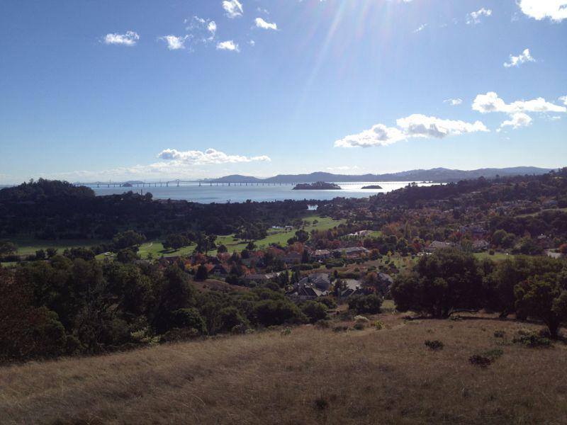 A panoramic view of a green landscape with residential areas, hills, and a body of water in the distance. The scene is bright and sunny, featuring a blue sky with scattered clouds and a bridge spanning the water. China Camp mountain bike trail.