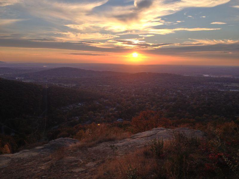 A scenic sunset view from a mountain overlook, showcasing vibrant orange and yellow hues in the sky while the sun sets over a valley filled with trees and distant hills, creating a tranquil and picturesque landscape. Blevins Gap Preserve mountain bike trail.