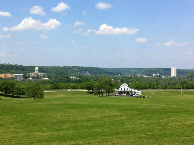 A vibrant green field stretches out under a clear blue sky dotted with a few clouds. In the foreground, a small white building with a porch is visible, surrounded by trees. In the background, rolling hills are present, along with structures that hint at a cityscape, featuring a dome-shaped building and a taller tower. The scene captures a mix of natural beauty and urban elements, showcasing a tranquil landscape. Capital View mountain bike trail.