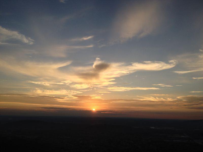 Sunset over a vast landscape, with the sun dipping below the horizon, casting warm hues of orange and gold across the sky. Wispy clouds reflect the fading light against a backdrop of soft blue and distant hills. The scene captures the tranquility of twilight in nature. Blevins Gap Preserve mountain bike trail.