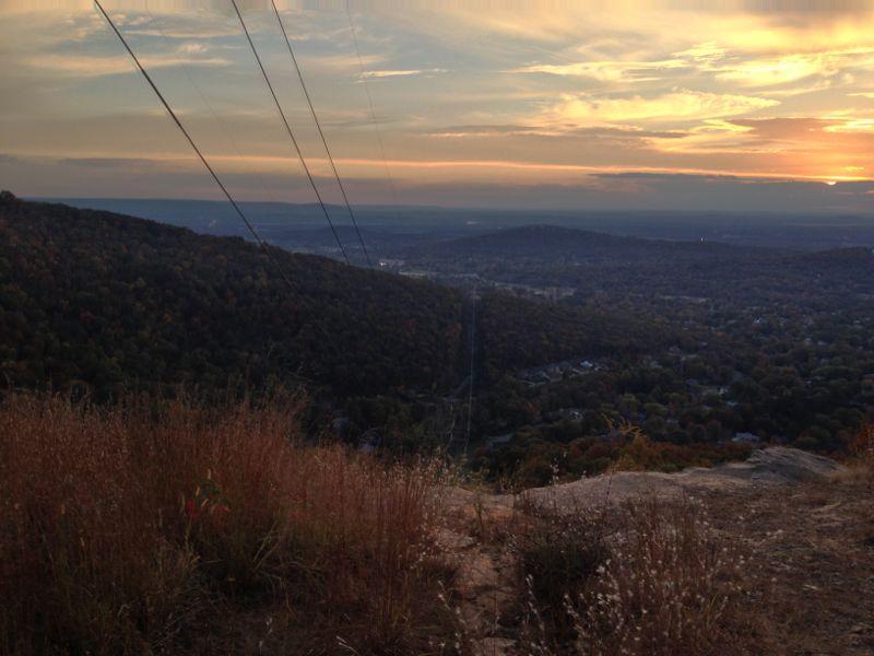 A panoramic view of rolling hills and valleys during sunset, showcasing a vibrant sky with hues of orange and yellow. In the foreground, rocky terrain with sparse grass and vegetation is visible, while power lines stretch across the scene. The landscape below features a mix of residential areas and wooded hills. Blevins Gap Preserve mountain bike trail.