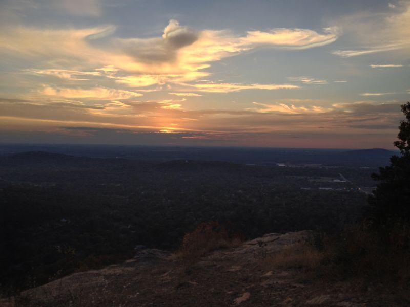 A scenic view of a sunset over a mountainous landscape, with swirling clouds illuminated in shades of orange, pink, and purple. The foreground shows rocky terrain and sparse vegetation, while the expansive valley below is blanketed in greenery. Blevins Gap Preserve mountain bike trail.