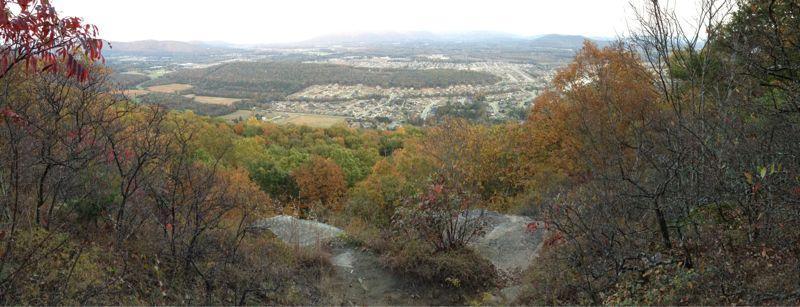 A panoramic view from a hillside showcasing a valley below, filled with a mix of green and autumn-colored trees, with distant mountains in the background. The scene captures the transition of seasons, reflecting vibrant fall foliage amidst a clearer landscape. Blevins Gap Preserve mountain bike trail.