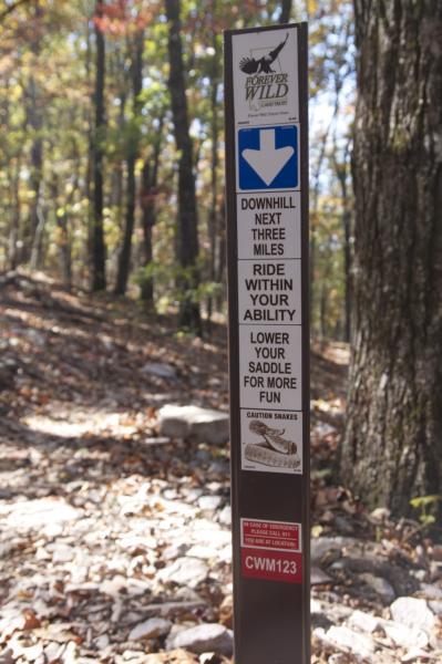 A trail marker indicating a downhill path for the next three miles, with safety instructions to ride within your ability and lower your saddle for more fun, alongside a caution about snakes. The sign is surrounded by a wooded area with fallen leaves on the ground. Coldwater Mountain mountain bike trail.