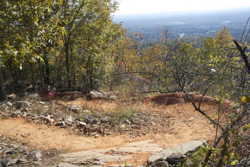 Winding dirt trail through a wooded area, leading down a rocky slope, with vibrant autumn foliage and a distant landscape visible under a clear sky. Coldwater Mountain mountain bike trail.