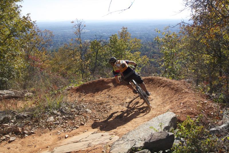 A cyclist navigating a dirt trail on a steep hillside, surrounded by trees displaying autumn foliage. In the background, a panoramic view of the landscape stretches out under a clear blue sky. The rider is leaning into a curve on the trail, showcasing dynamic movement and skill. Coldwater Mountain mountain bike trail.