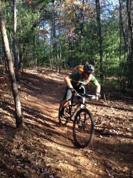 A mountain biker navigating a winding dirt trail through a wooded area, with trees lining the path and autumn leaves on the ground. The rider is leaning forward, demonstrating speed and agility, while wearing a helmet and a green shirt. Sunlight filters through the trees, creating a vibrant outdoor scene. Jake to Bull Mountain Connecter mountain bike trail.