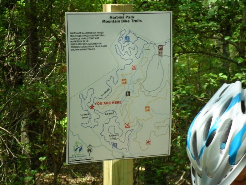 Map of Harbins Park Mountain Bike Trails, showing various bike paths and trails, with a marked location indicating "You Are Here," and a biking helmet resting against a wooden post in a wooded area. Harbins Park mountain bike trail.
