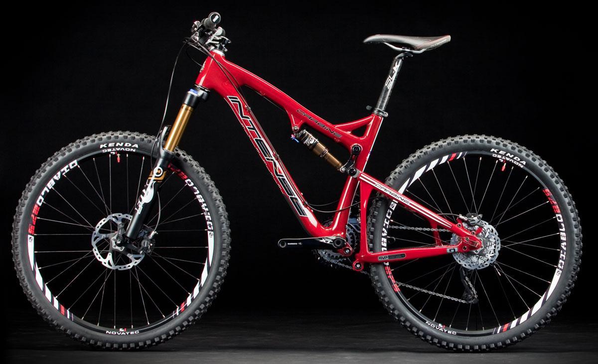 Intense Tracer 275: A red mountain bike with a glossy finish, featuring a full suspension design, large knobby tires, and prominent branding on the frame and wheels. The bike is showcased against a black backdrop, highlighting its sleek lines and modern components.