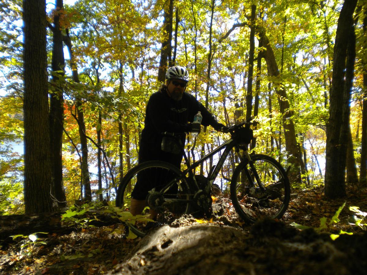A mountain biker standing next to a bike on a trail in a forest during autumn, surrounded by colorful leaves and sunlight filtering through the trees. The biker is wearing a helmet and holding a water bottle. Lick Creek Trail Short loop mountain bike trail.