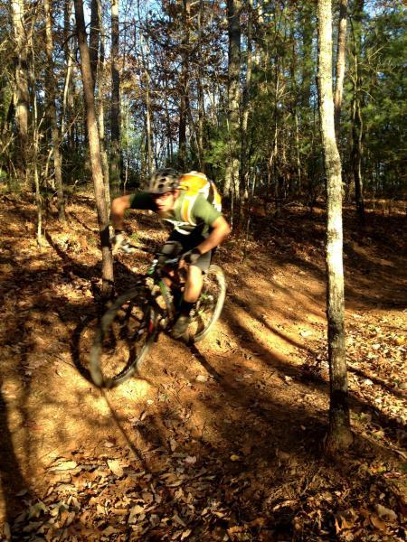 A cyclist navigating a winding trail through a wooded area, surrounded by trees and fallen leaves. The rider leans into a turn, wearing a helmet and a backpack, with sunlight filtering through the foliage. Jake to Bull Mountain Connecter mountain bike trail.