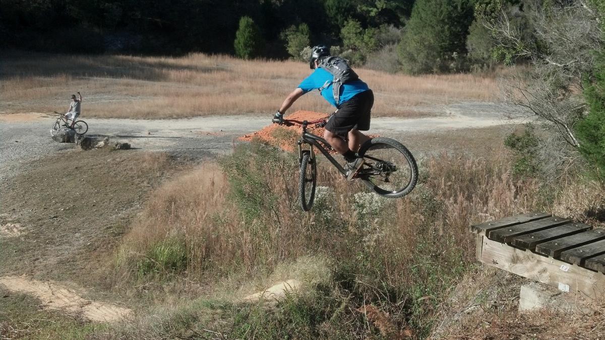 A mountain biker in a blue shirt riding off a wooden ramp, airborne above a dirt path and surrounded by tall grass and sparse vegetation. Another cyclist can be seen in the background, standing beside their bike. Santos mountain bike trail.