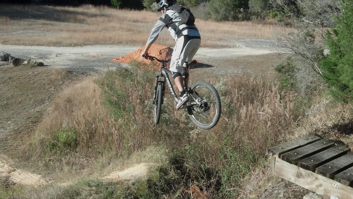 A mountain biker performing a jump over a small drop-off, surrounded by tall grass and a dirt path, with a pile of dirt in the background. The rider is wearing protective gear and is airborne above the ground. Santos mountain bike trail.