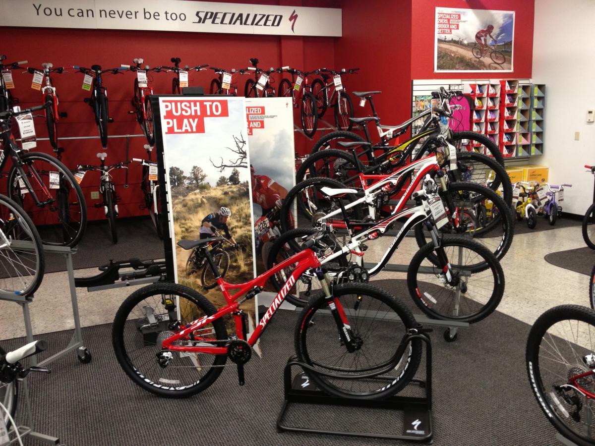 A display of various bicycles in a Specialized bike shop, featuring a prominent red mountain bike in the foreground. The background includes several models hanging on a wall with price tags, along with a promotional poster encouraging outdoor activity. The shop has a red and white color scheme with a carpeted floor.