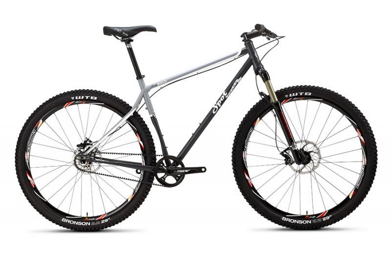 SPOT Honey Badger Singlespeed: A modern mountain bike featuring a sleek gray and black frame, wide tires with a rugged tread pattern, and disc brakes. The bike is designed for off-road cycling, showcasing a rigid fork and a single gear setup, ideal for trails and rough terrains.