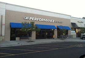 storefront of a Performance Bicycle shop, featuring a beige exterior, blue awnings, and large windows displaying bicycles and cycling gear.