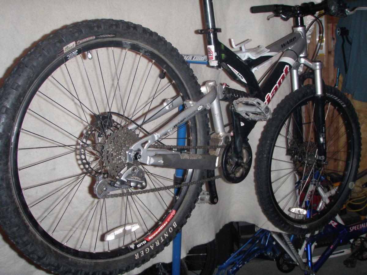 Giant Warp DS3: A mountain bike hanging on a wall, showcasing its silver frame, black tires, and various components such as the chain, gears, and pedals. The background consists of a plain surface with some tools and bike parts visible.