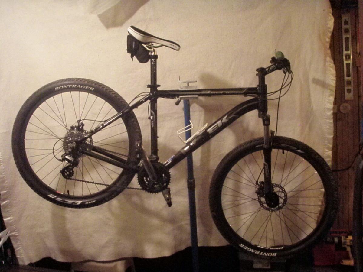 Trek 3700: A black mountain bike is displayed on a repair stand against a light-colored backdrop. The bike features Bontrager tires and a standard mountain bike frame design, with visible gears and brakes. The seat is positioned above the frame, and the handlebars are turned slightly to the side.