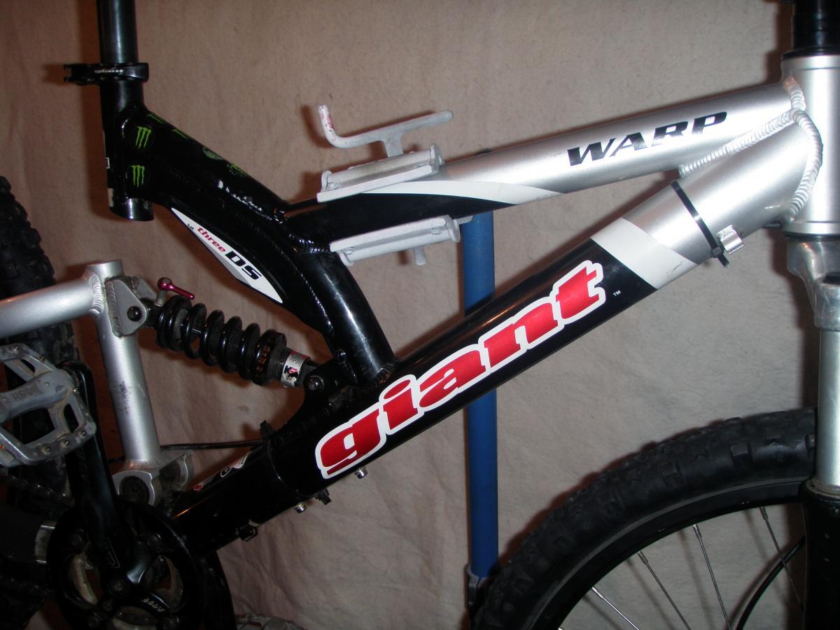 Giant Warp DS3: Close-up of a mountain bike frame featuring a combination of black and silver colors. The frame is branded with the logos "Giant" and "Warp." The image highlights the bike's suspension system, including a prominent coil spring, and shows parts of the drivetrain and wheels. The background is neutral, emphasizing the bike components.