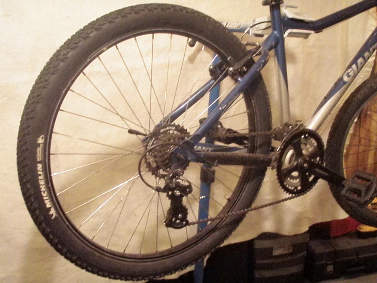 Michelin XCR Dry2: Alt text: A close-up view of the rear wheel and drivetrain of a blue mountain bike, showing the gear mechanism, chain, and tire with a visible logo. The background is a neutral wall with storage boxes.