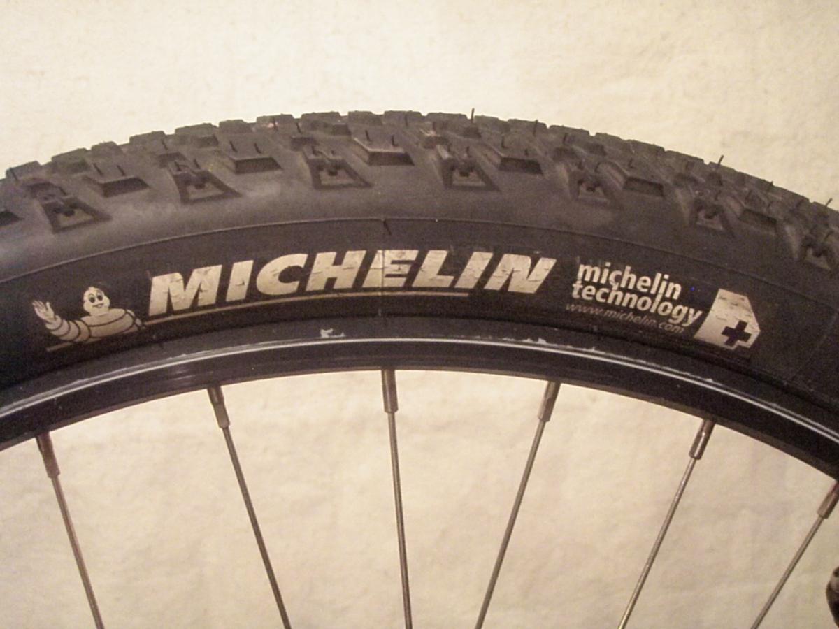 Michelin XCR Dry2: Close-up of a Michelin bicycle tire showcasing its tread pattern, branding, and the Michelin logo, featuring the iconic mascot. The image highlights details like the text "michelin technology" and the web address in a simple background.