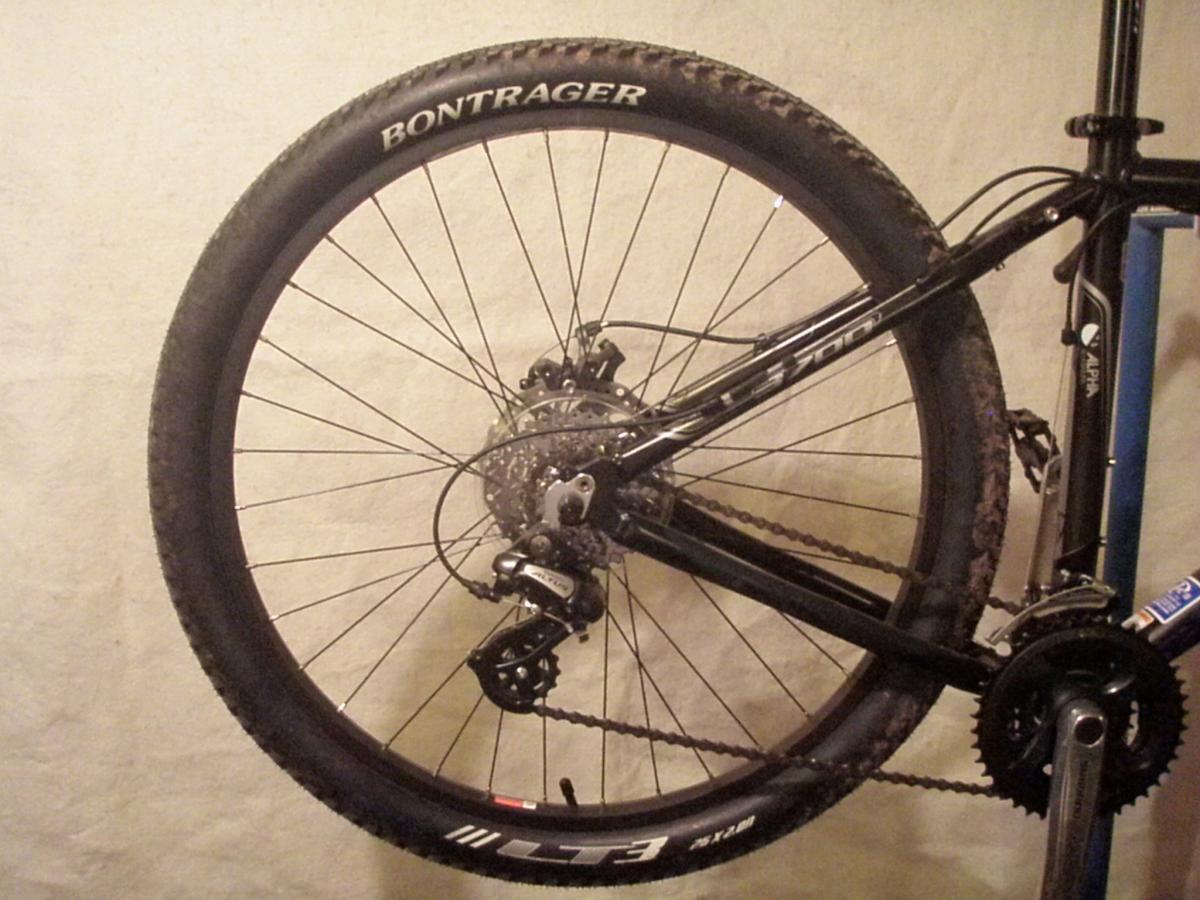 Bontrager LT3: A close-up image of a mountain bike's rear wheel, showcasing the Bontrager tire, chain, and derailleur. The bike is hung against a plain background, with visible dirt on the tire, indicating recent use.