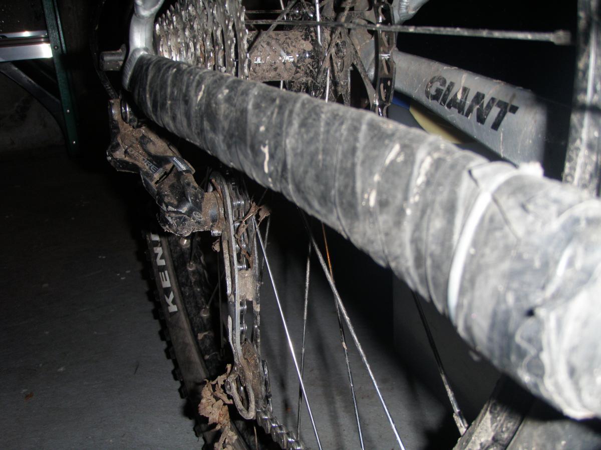 Specialized Specialized Specialized Standard Tube 26"x1.5/2.2 (Presta): Close-up view of a mountain bike's rear gear system, showcasing a dirty chain and derailleur. The bike frame, partially visible, has the brand name "Giant." The background includes a blurred green object and a concrete floor.