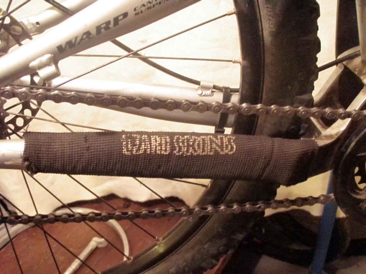 Lizard Skins Jumbo Chain Stay Guard: Close-up image of a bicycle with a focus on the chain and protective wrapping on the chainstay. The bike frame is silver, labeled "WARP," and the protective wrap reads "LIZARD SKINS." The background shows a blurred indoor setting with a wooden floor.