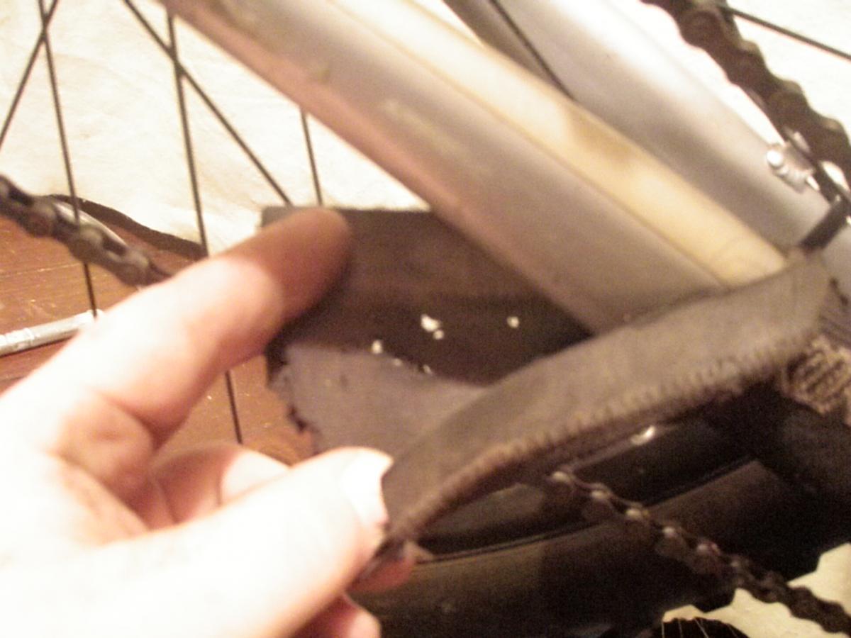 Lizard Skins Jumbo Chain Stay Guard: A close-up image of a hand pointing to a frayed piece of fabric attached to a bicycle frame, with bicycle chain and part of the wheel visible in the background. The surface is wooden, and there are tools laid out nearby.
