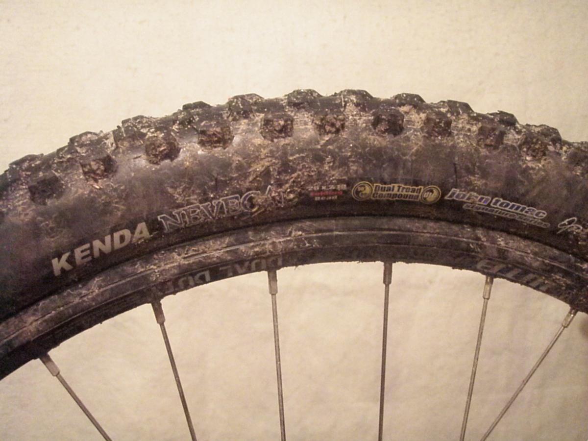Kenda Nevegal: Close-up view of a mountain bike tire with a rugged tread pattern, partially covered in dirt. The tire is labeled "Kenda Nevegal" and shows features like "Dual Tread Compound." The rim and spokes are visible in the background.