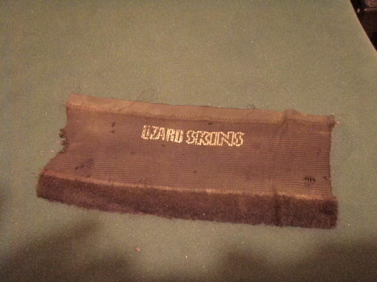Lizard Skins Jumbo Chain Stay Guard: A piece of fabric with the text "LIZARD SKINS" embroidered in metallic thread, laying on a green surface. The fabric has frayed edges and some dirt marks, suggesting it may have been used previously.