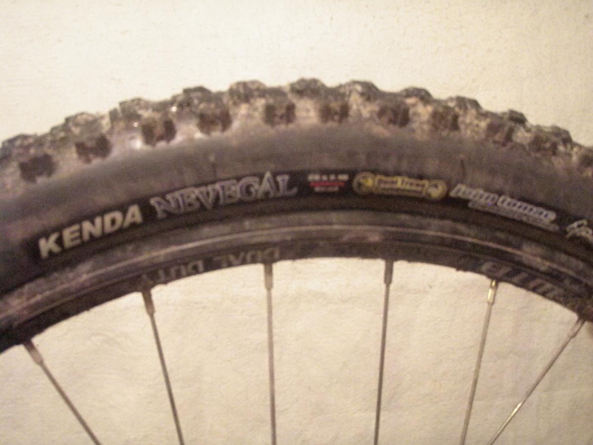 Kenda Nevegal: Close-up view of a Kenda Nevegal mountain bike tire, showcasing the tread pattern and sidewall branding, with a slightly worn appearance. The background is a plain, light-colored wall.