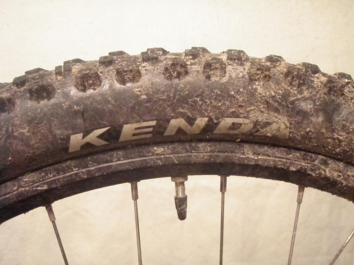 Kenda Nevegal: Close-up image of a dirty Kenda mountain bike tire, showcasing the textured tread and rim. The tire is partially muddy, indicating off-road use.