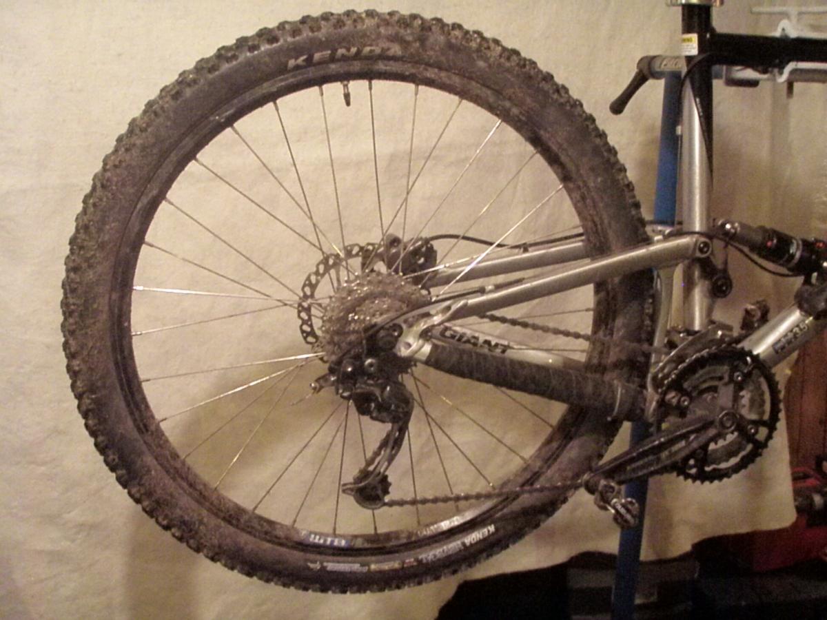 Kenda Nevegal: A close-up view of a mountain bike's rear wheel and drivetrain, showing a dirty tire labeled "KENDA" and various gears and components of the bike. The bike is mounted on a rack against a neutral background.