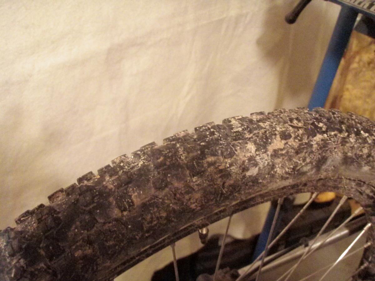 Kenda Nevegal: Close-up of a bicycle tire with a worn tread pattern, featuring dirt and mud residue, set against a plain background.