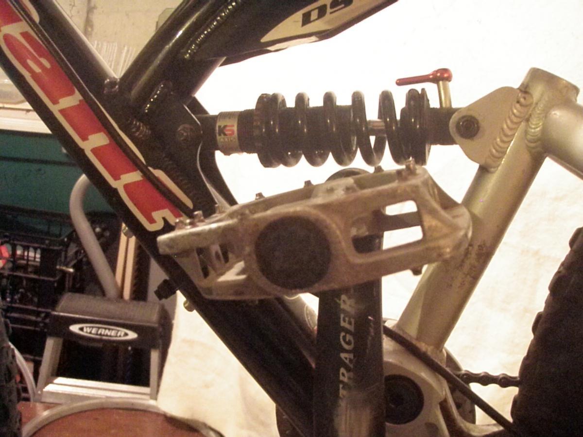 Close-up image of a bicycle's drivetrain, showing the crankset, pedal, and rear suspension system. The bicycle frame features visible branding, and there is a coil spring shock absorber integrated into the rear suspension. The background is slightly blurred, focusing attention on the mechanical components.