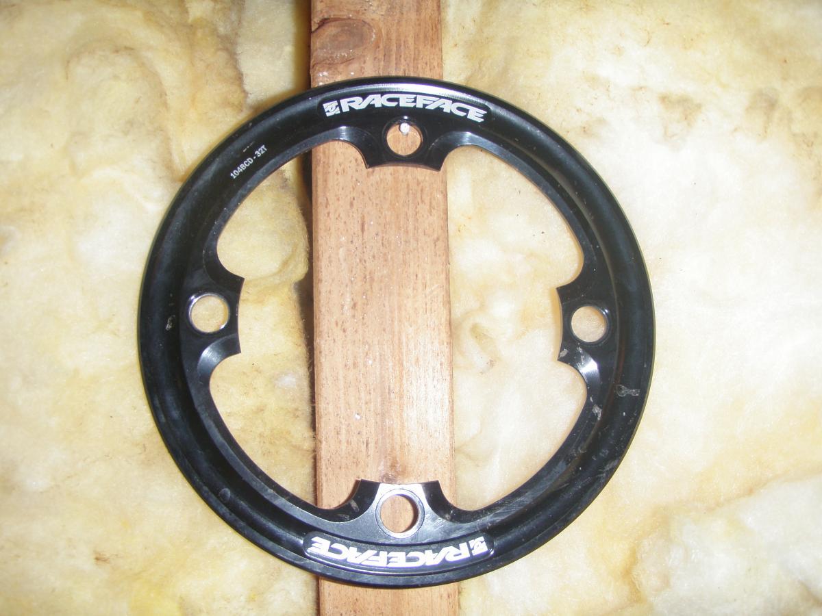 Race Face Lightweight Bash: Black bicycle chainring with "Raceface" branding, positioned on a wooden surface against a background of insulation material. It features cutouts and holes for attachment.