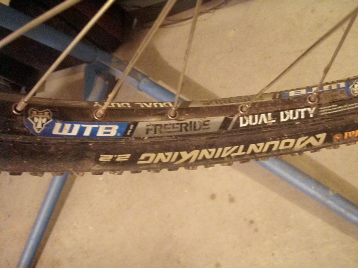 Continental Mountain King II: A close-up image of a mountain bike tire showcasing the WTB FreeRide Dual Duty branding. The tire has a rugged tread and is positioned on a bike stand, with some dirt visible on the surface. The background is a plain garage floor.