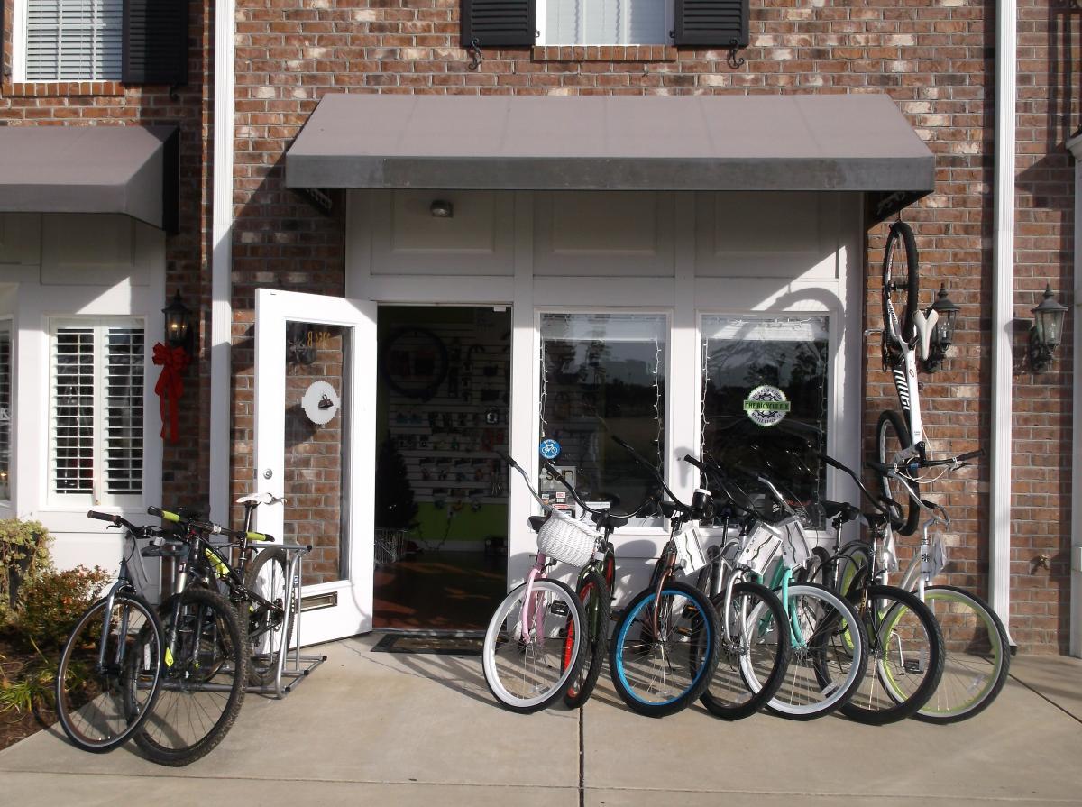 A storefront with a variety of bicycles lined up outside, featuring different colors and styles. The entrance has a wreath decoration, and there are windows displaying bike accessories in the background. The building has a brick facade and a dark awning above the door.
