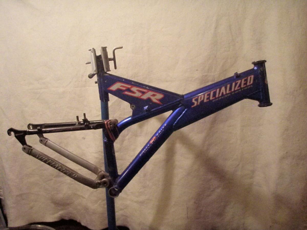 Specialized FSR: A blue mountain bike frame with a highlighted logo that reads "Specialized" and "FSR," mounted on a bike repair stand. The frame features dual suspension components, with one arm in black and another in gray. The background is a plain light-colored fabric.