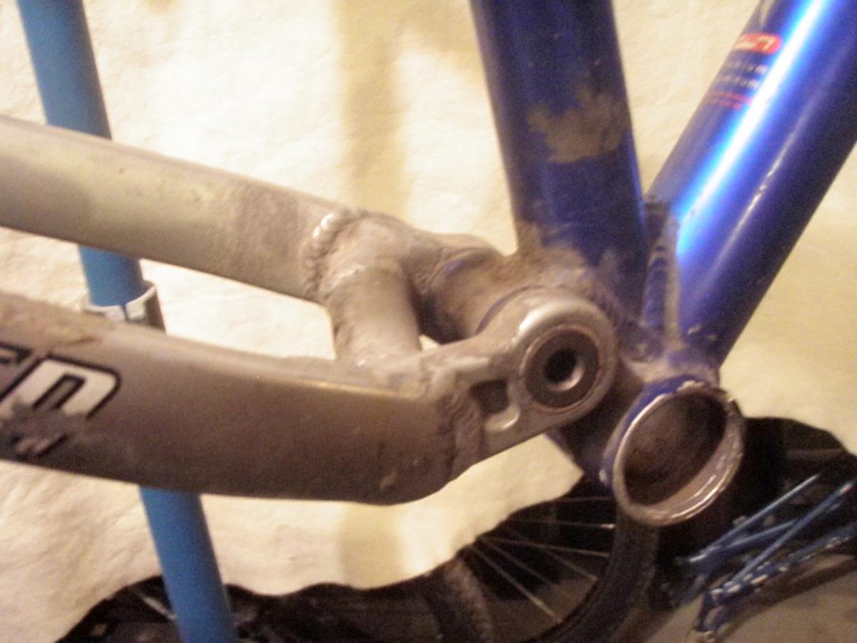 Specialized FSR: Close-up view of a bicycle frame showing the connection between the crank arm and the bottom bracket. The frame is primarily blue with some visible dirt and wear. A part of the bicycle stand is visible in the background, along with a hint of a wheel.