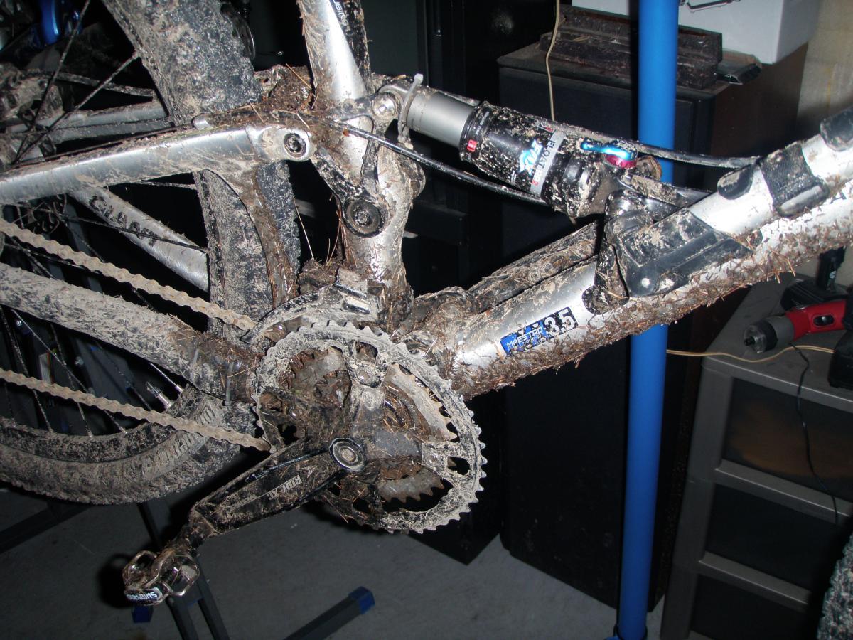 Giant Anthem 2: Close-up image of a muddy mountain bike's drivetrain and rear suspension, showing significant dirt accumulation on the chain, gears, and frame components. The bike is positioned on a repair stand, highlighting the wear and tear from off-road riding.