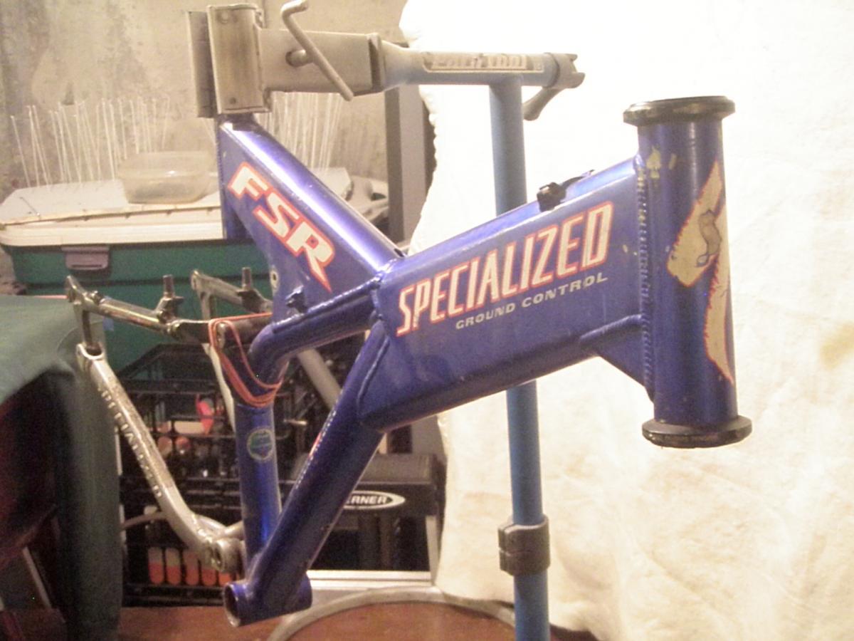 Specialized FSR: Alt text: Close-up of a blue Specialized FSR bicycle frame displayed on a repair stand, with the "Ground Control" label visible. The background shows blurred objects, indicating a workshop or storage area.