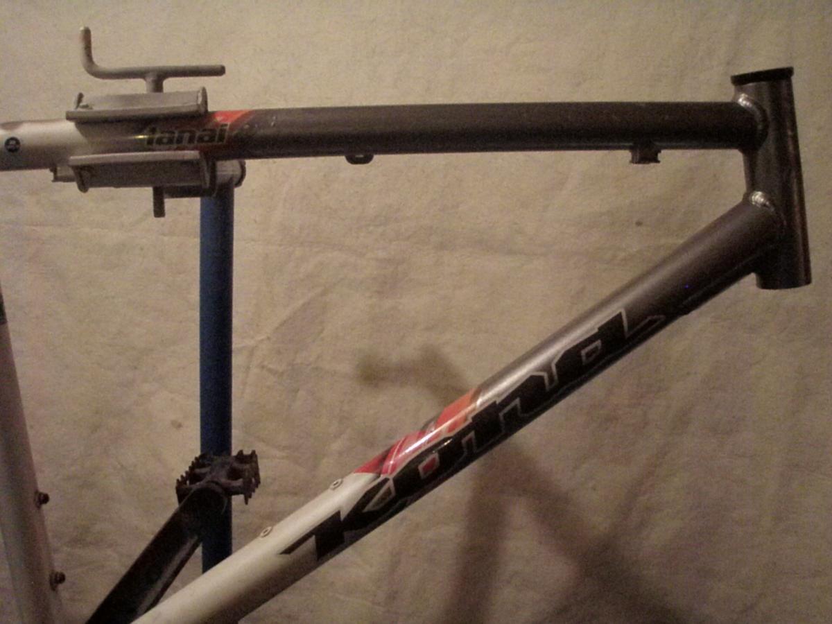 Kona Lana'i: Image of a bicycle frame, showing two sections of the frame: the top tube and the down tube, which are partially connected. The frame features a combination of gray and black colors with visible branding. The frame is mounted on a blue bicycle stand against a neutral background.