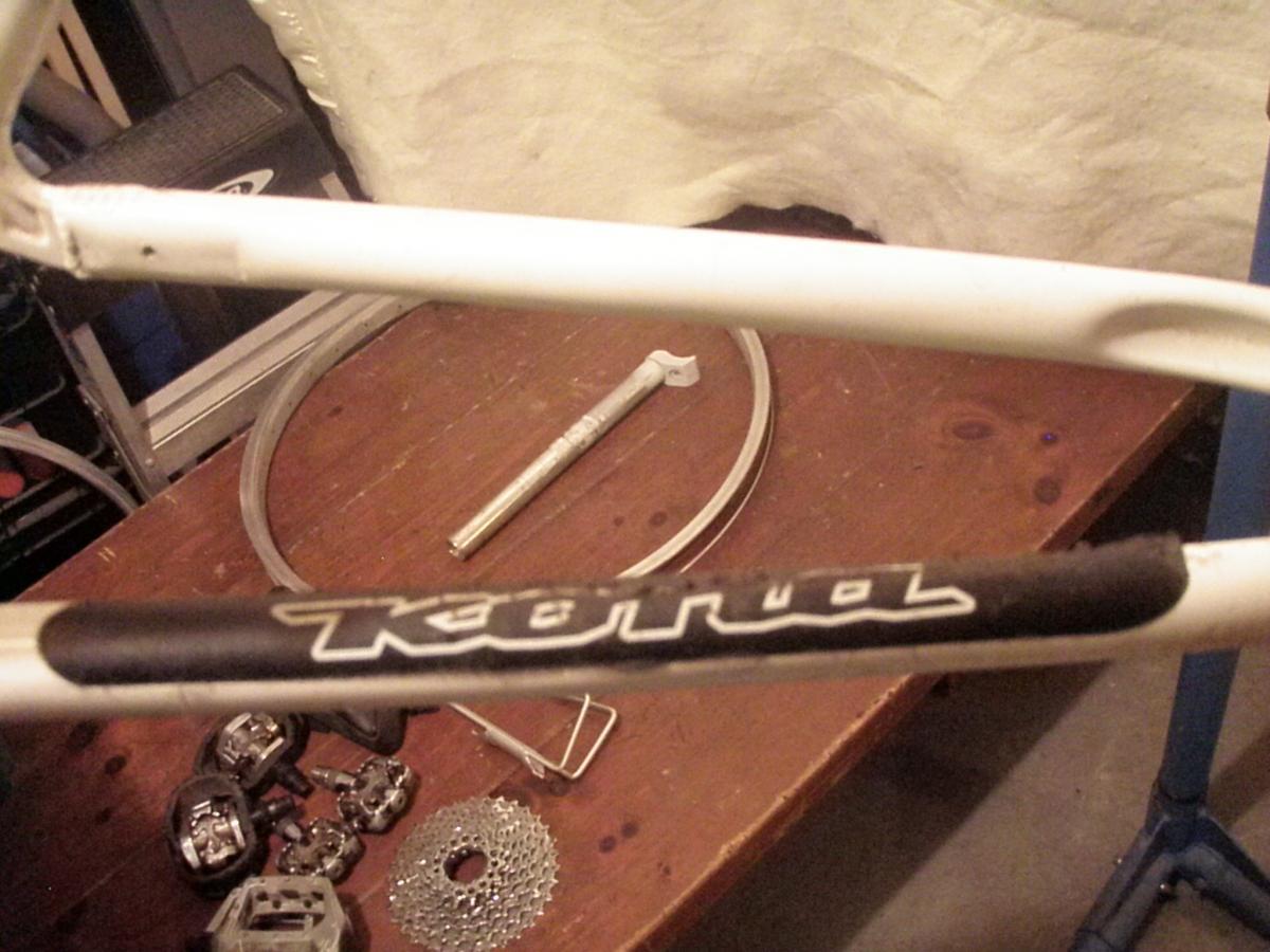 Kona Lana'i: Image of a bicycle frame with visible brand markings. The frame is predominantly white with a black section displaying the name "Kona." In the background, there are various bike components, including pedals, a gear cassette, and a tool, on a wooden surface.