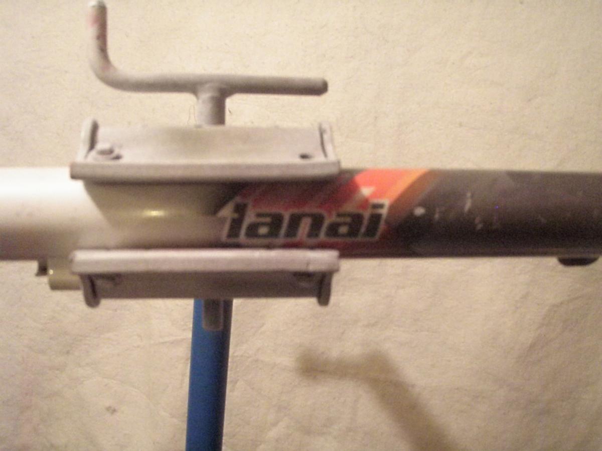 Kona Lana'i: Close-up image of a bike frame with a tanai logo, attached to a bike repair stand. The frame shows some wear and has a combination of white and dark colors. The background is a plain, light-colored wall.