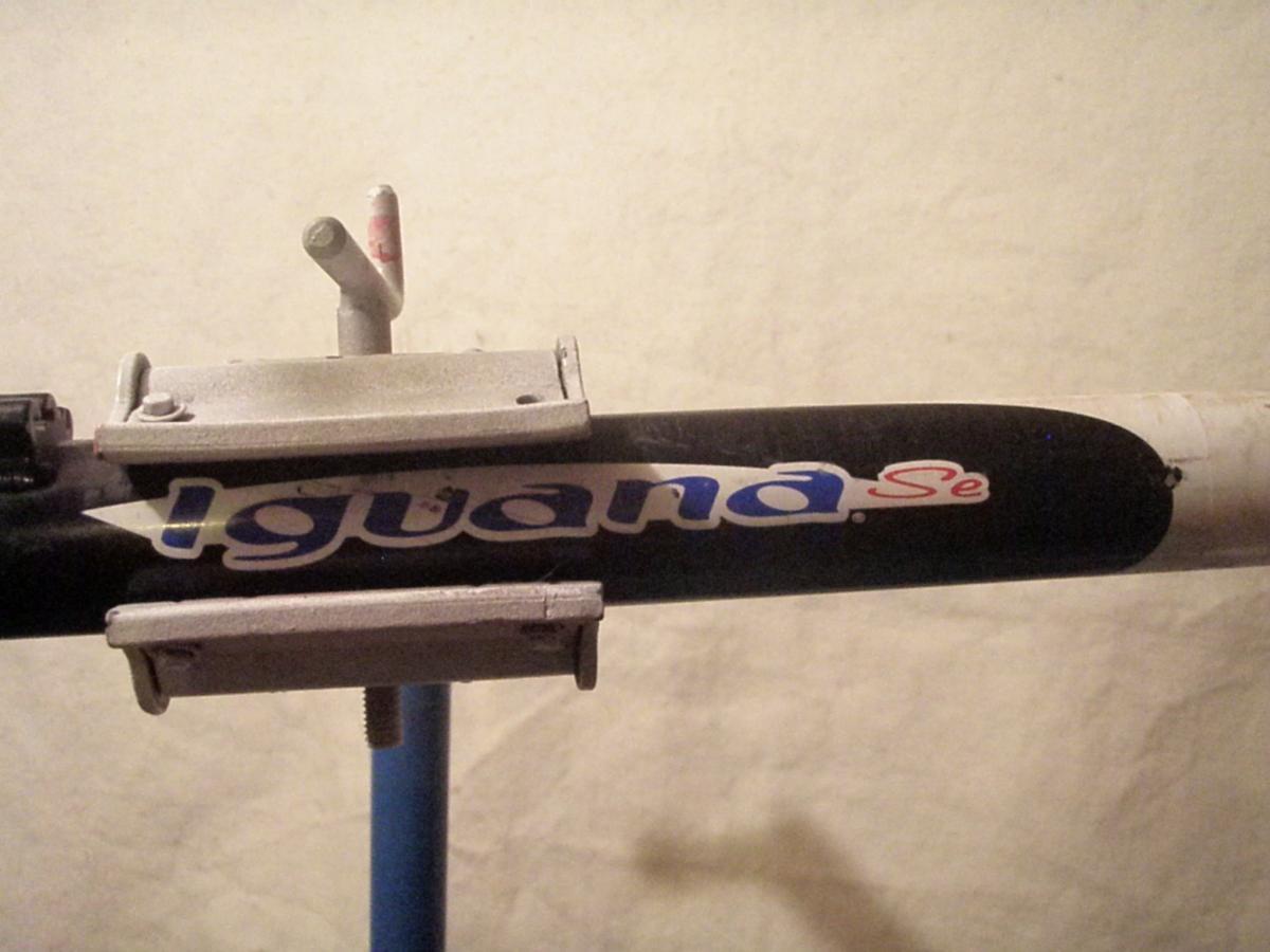 Giant Iguana: Close-up view of a bicycle frame with an "Iguana Se" logo. The frame is primarily black with a white section, and metal clamps are visible securing part of the frame. The background is a plain, light-colored wall.